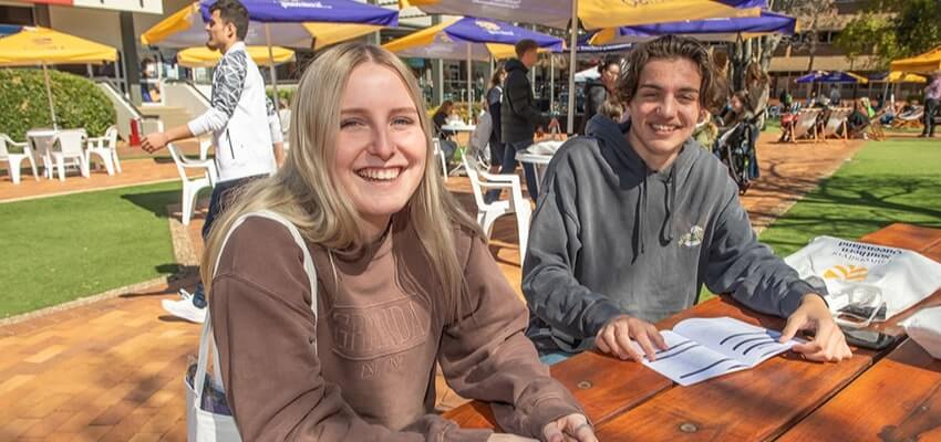 Two people sitting at a wooden picnic table outdoors, smiling. There are open papers in front of them and other people in the background.