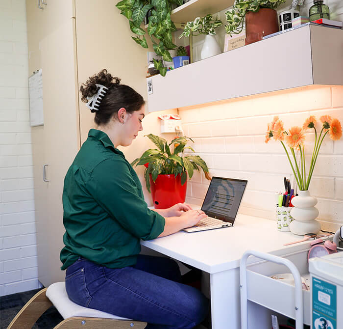 A woman in a green shirt sits at a white desk working on a laptop. The desk has plants and flowers, and there is a shelf above with more plants.