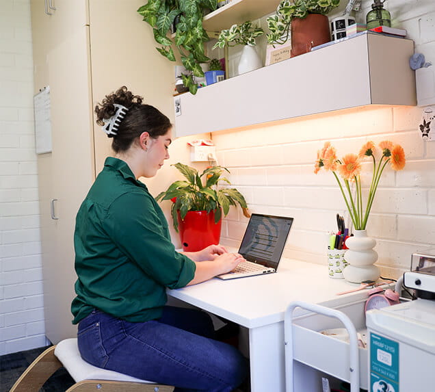 Woman in green shirt works on a laptop at a desk with plants, flowers, and shelves above her in a well-lit room.