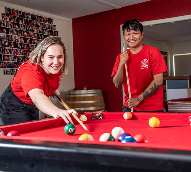 Two people wearing red shirts play pool on a red table. One person lines up a shot while the other watches. A photo collage is on the wall in the background.