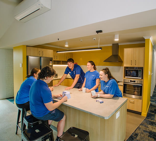 Five people in blue shirts sit and stand around a kitchen island, engaging in conversation.