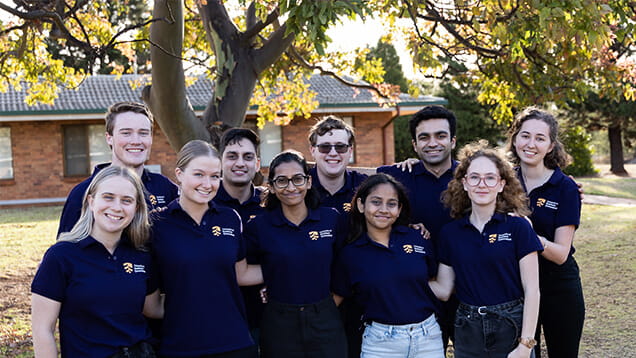 Group of ten people wearing matching navy polo shirts, standing outside under a tree with a building in the background.