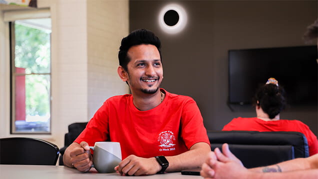 A person in a red shirt smiles while holding a mug indoors. A partial solar eclipse is visible through the window.