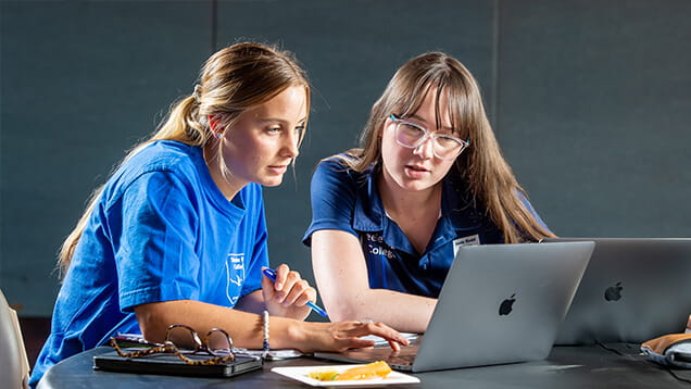 Two people sitting at a table, focusing on a laptop screen. They are wearing glasses. A notebook, pen, and a plate with a snack are also on the table.