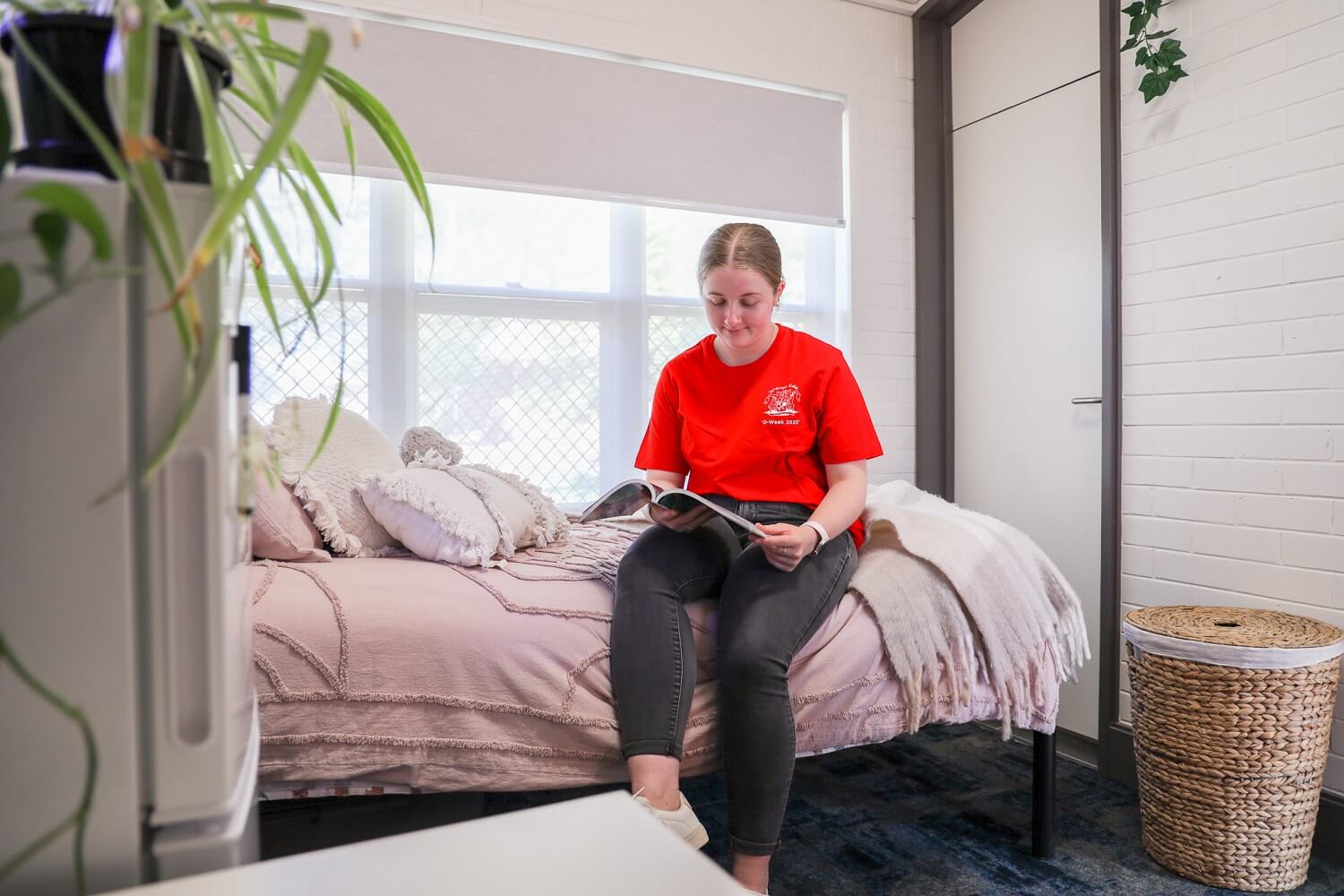 Person in a red t-shirt sits on a bed reading a book in a bright room with white walls. A plant is visible on the left, and a woven basket is on the floor.