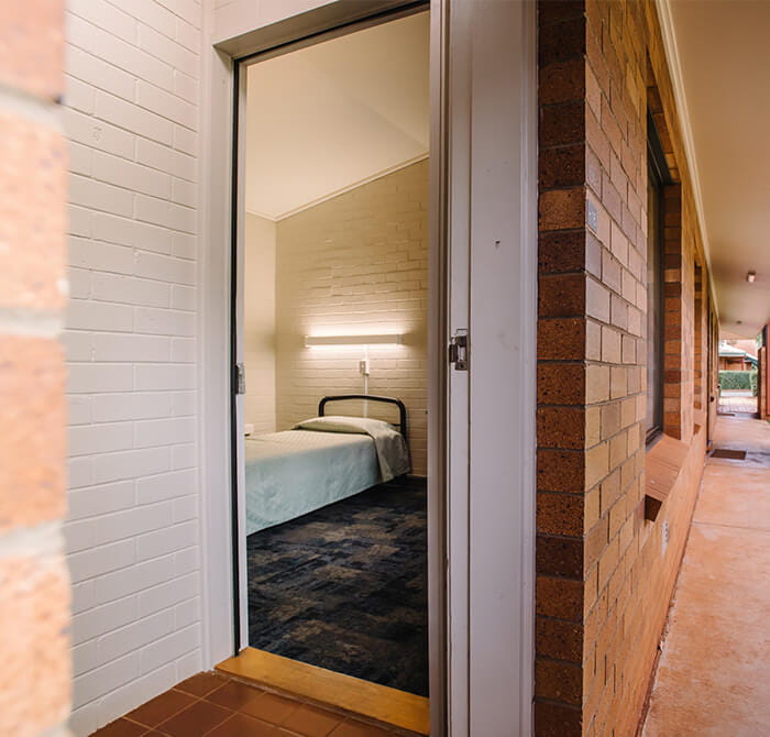 Open door view into a simple room with a single bed, white walls, and a glowing light fixture on the wall.