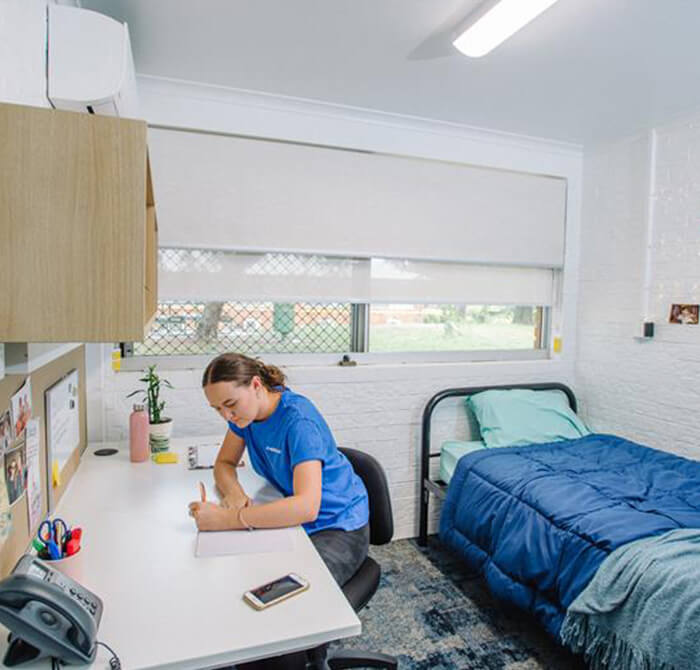 Person sitting at a desk writing in a notebook. The room has a bed with a blue blanket, a window with a blind, and shelves with various items.