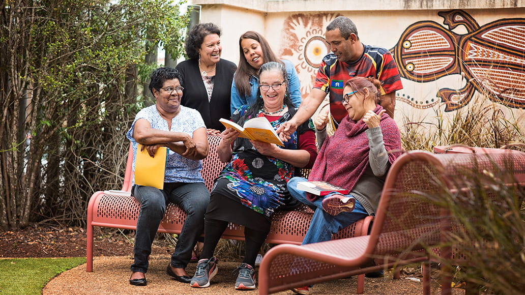 A group of people sitting on benches outside, smiling and reading books. There is a mural on the wall behind them.