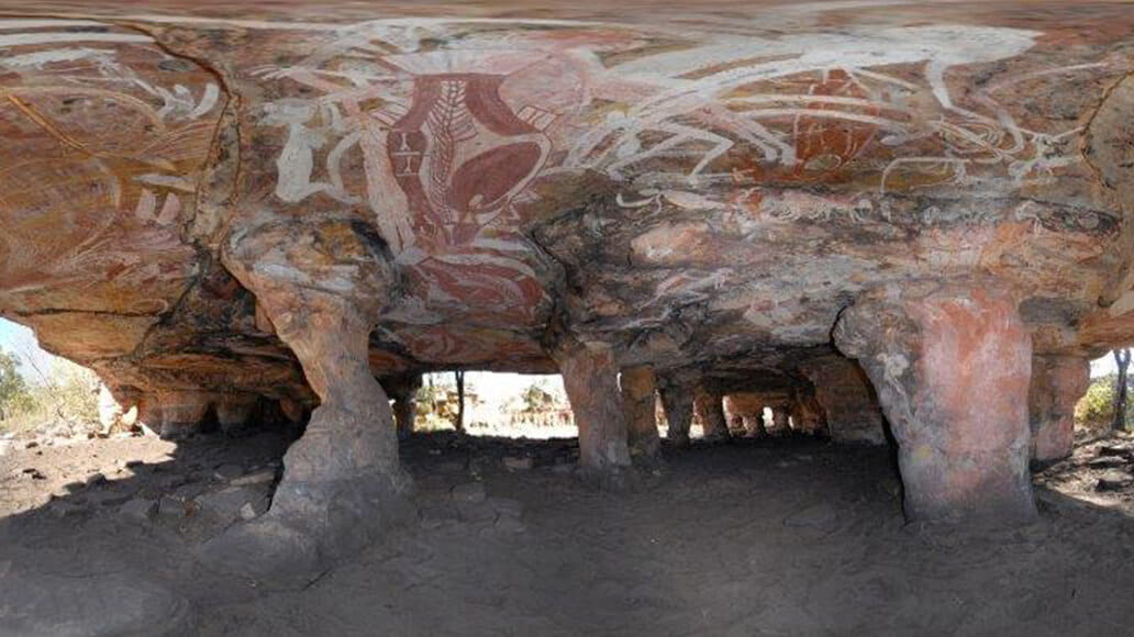 Rock formation with columns and painted indigenous art on the ceiling in a cave setting.