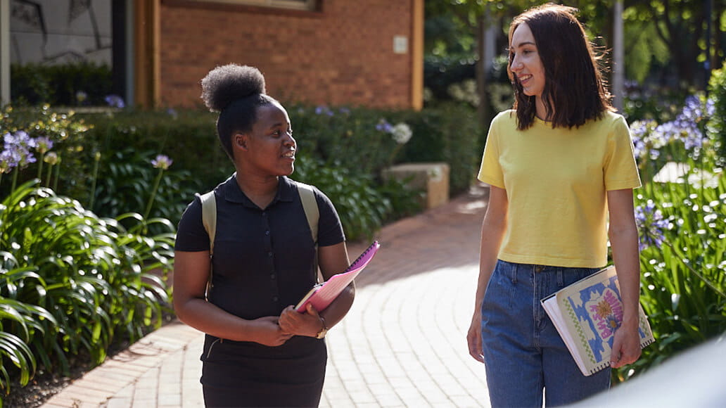 Two students walk and converse on a brick path lined with plants. One holds a pink folder, and the other carries a floral-patterned binder.