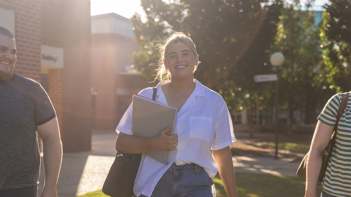 A person holding a notebook and smiling while walking outside with two others in a sunlit area.