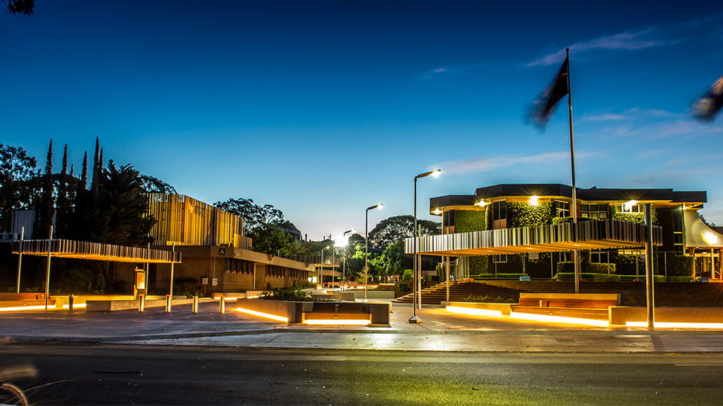 Building with sleek modern architecture and a flagpole at dusk. Pathways are illuminated with soft yellow lighting, trees and vegetation in the background. Clear sky, light fading.