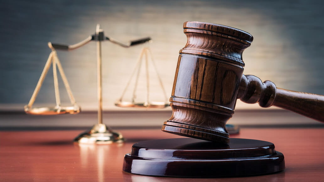 A wooden gavel rests on a sound block in the foreground, with a set of gold balance scales in the background on a wooden surface.