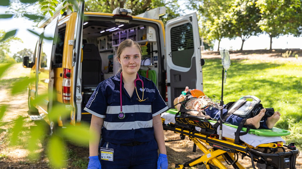 Paramedic standing next to an open ambulance with a patient on a stretcher in a park setting.