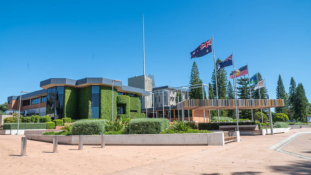 A modern government building with green ivy walls, multiple flags flying, and surrounding greenery under a clear blue sky.