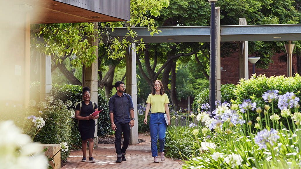 Three people walking on a brick path surrounded by greenery and flowers in a landscaped garden. They appear to be engaged in conversation.