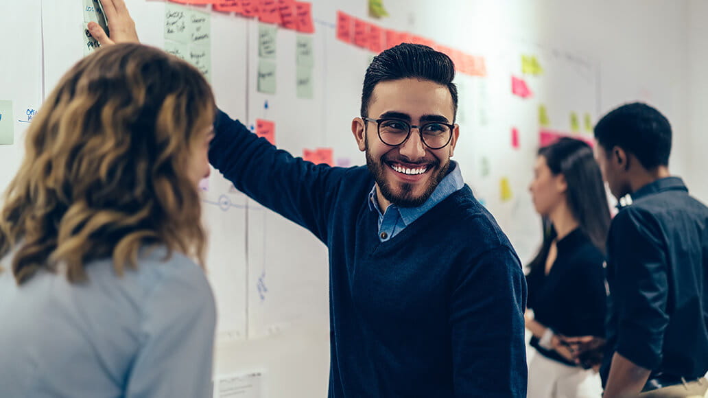 A man in glasses and a blue sweater smiles while pointing to a whiteboard in a collaborative office setting with three colleagues.