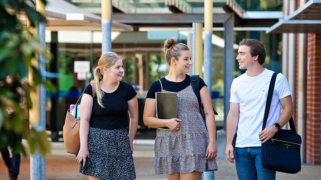 Three students walk outside, holding books and bags, conversing and smiling.