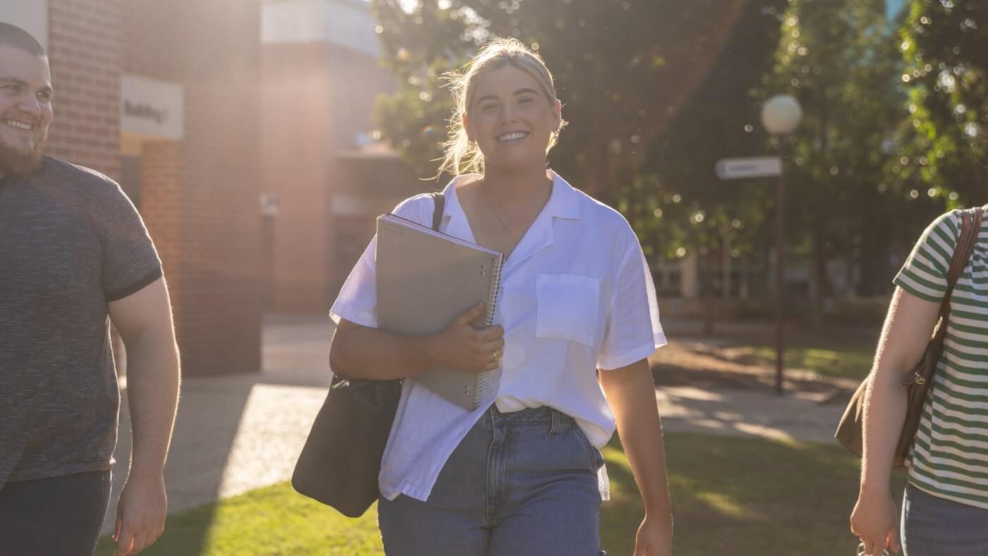 A person in a white shirt and jeans walks outdoors holding a binder, flanked by two others, with sunlight in the background.