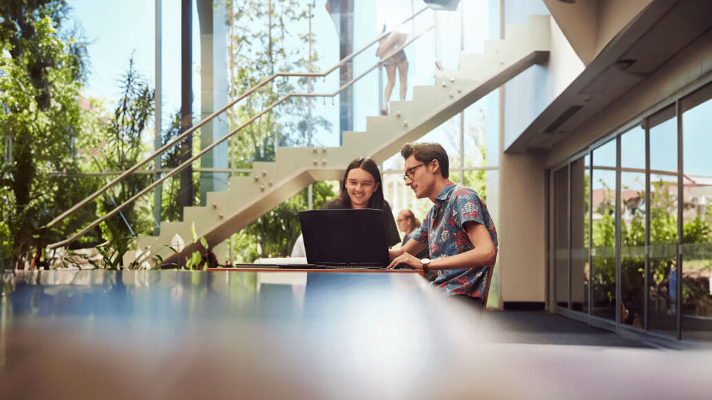 Two university students sit at table looking at a laptop smiling.