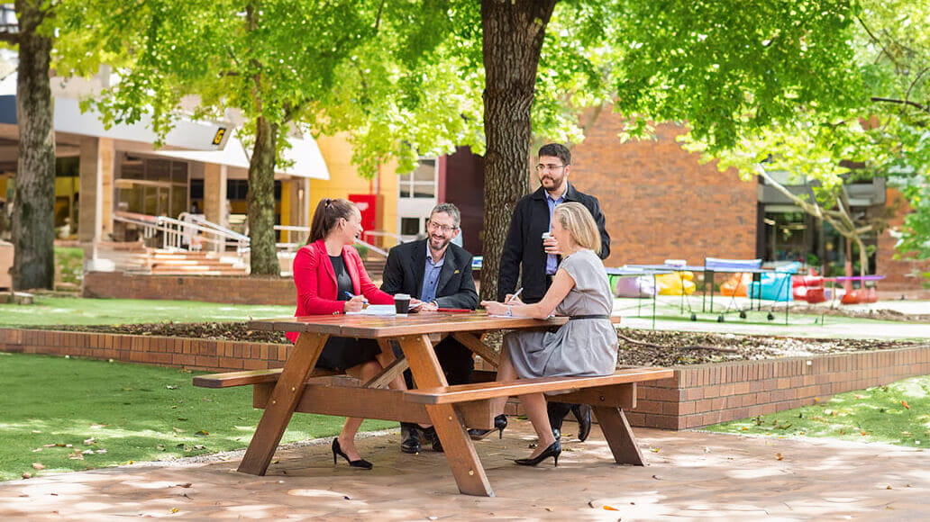 Four adults are seated at a wooden picnic table under trees in an outdoor area, engaged in conversation. One person stands nearby, observing. They appear to be in a park or campus setting.