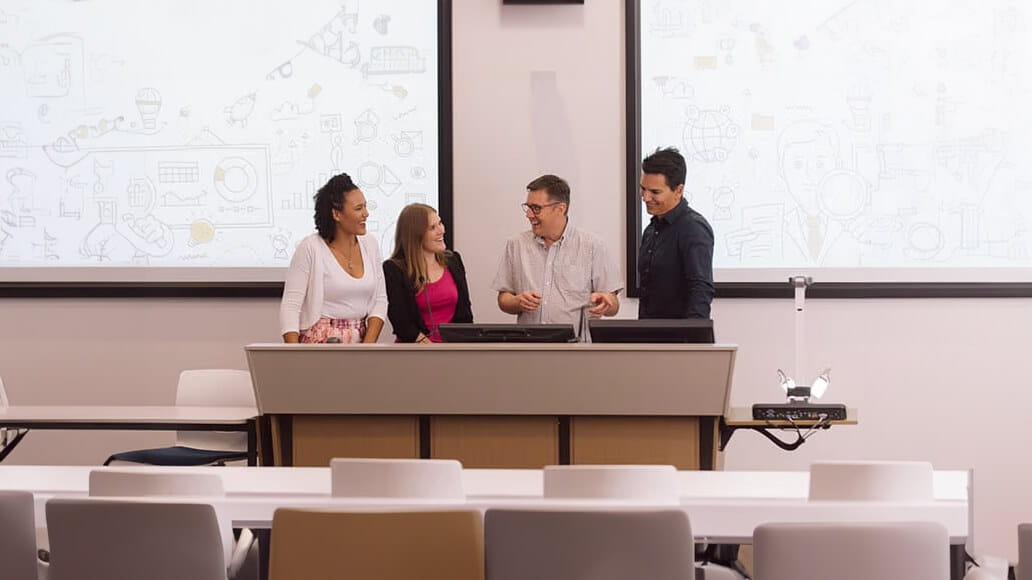 Four people stand and converse behind a lectern in a classroom with illustrated whiteboards in the background.