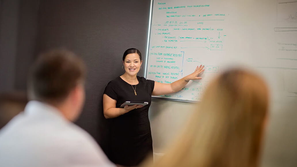 A woman stands near a whiteboard with mathematical equations, holding a tablet and gesturing while speaking to the audience. People are seated facing her, listening attentively.