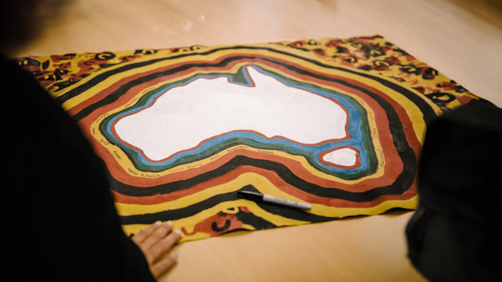 A person on the left is leaning over a hand-painted artwork depicting the map of Australia, framed by concentric stripes of red, yellow, brown, and black. A marker lies on the artwork.