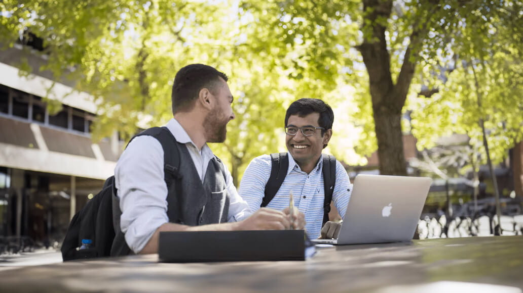 Two men sit at an outdoor table, talking and smiling, with a laptop open in front of them. They both have backpacks, and the background shows trees and a building.