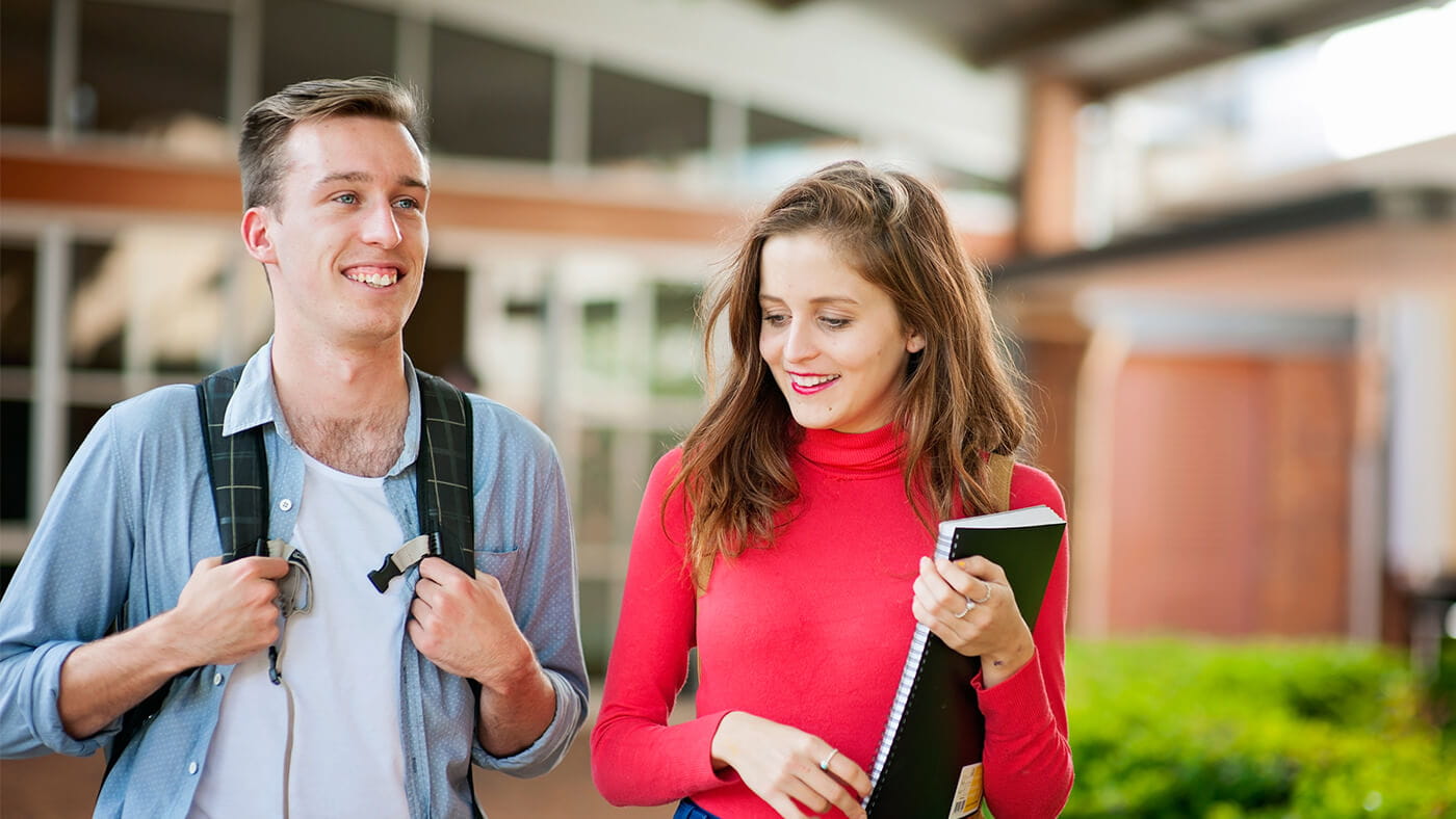 Two young adults, one wearing a backpack and the other holding notebooks, walk together outdoors, smiling and conversing. They appear to be on a college campus.