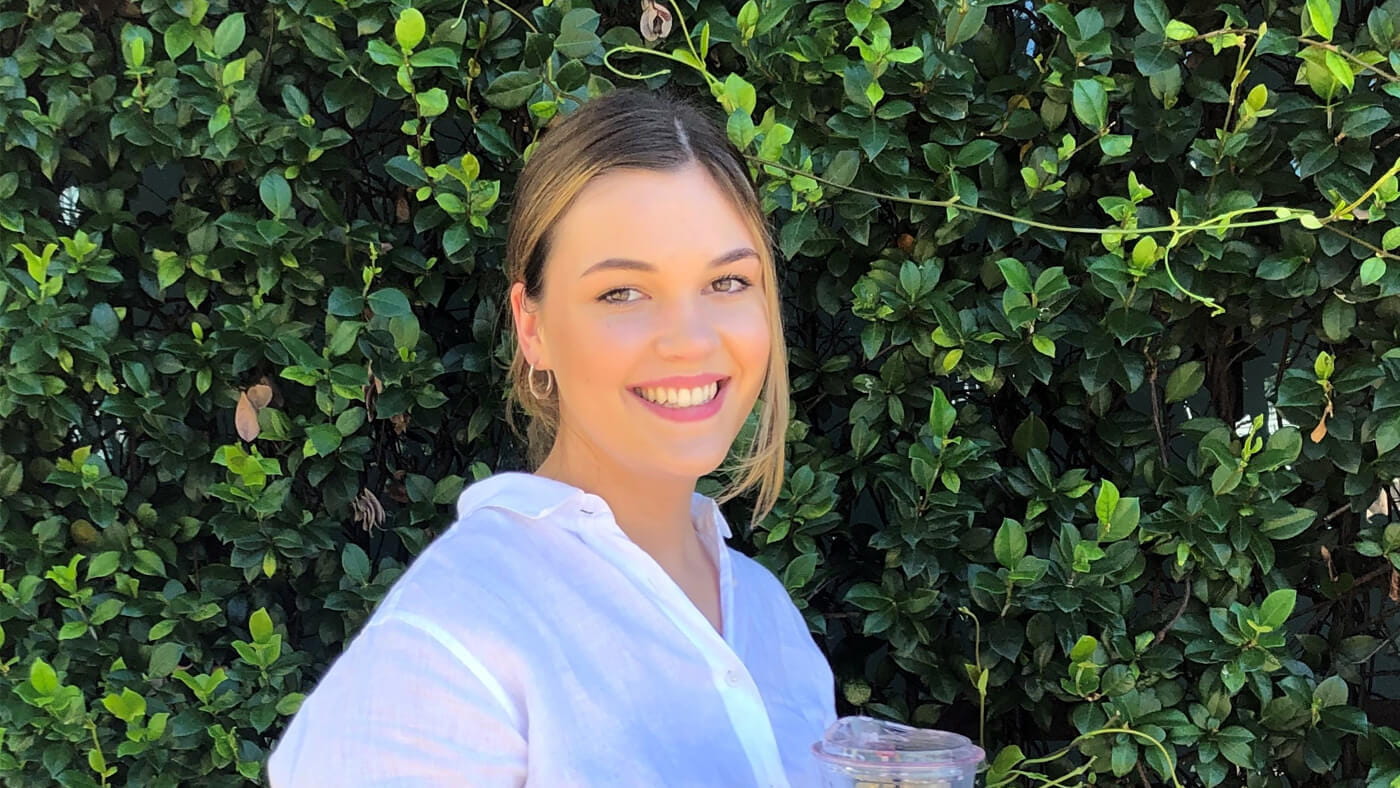 A woman in a light-colored blouse stands in front of a lush green hedge, smiling while holding a drink container.