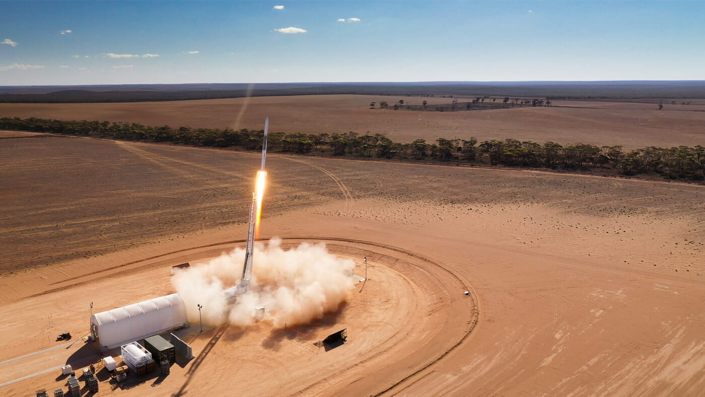 A rocket is launching from a desert landscape with a clear sky overhead. Structures and vehicles are situated near the launch pad, and a trail of smoke follows the ascending rocket.