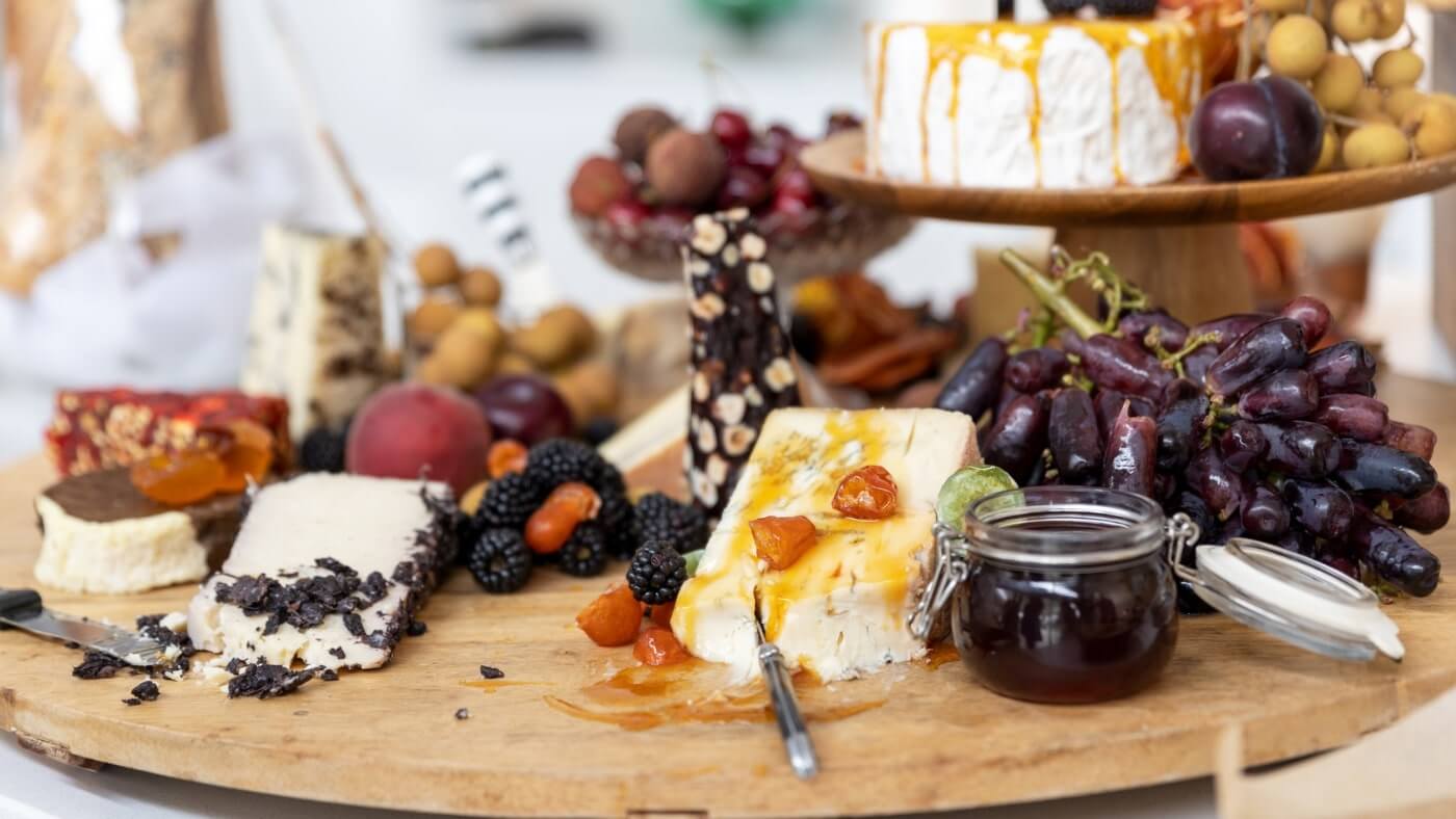 A wooden platter with assorted cheeses, fruit, and a jar of jam, featuring grapes, blackberries, and nuts.