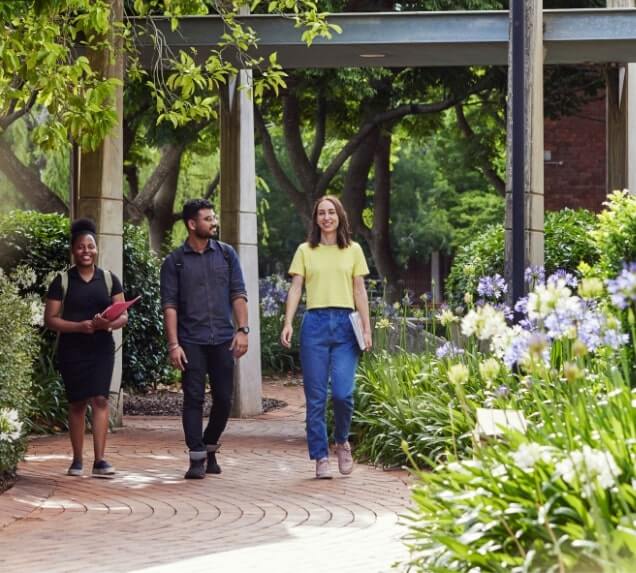 Three people walk on a garden pathway lined with flowers and trees at UniSQ Toowoomba.