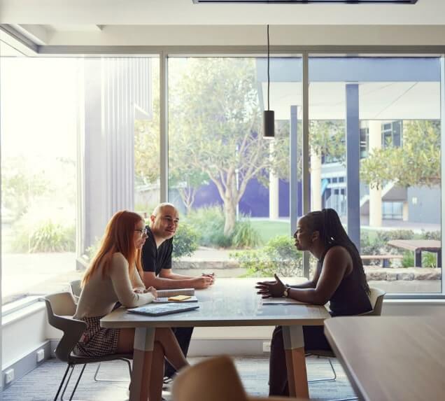 Three people sitting at a table in a bright room with large windows, engaged in conversation.