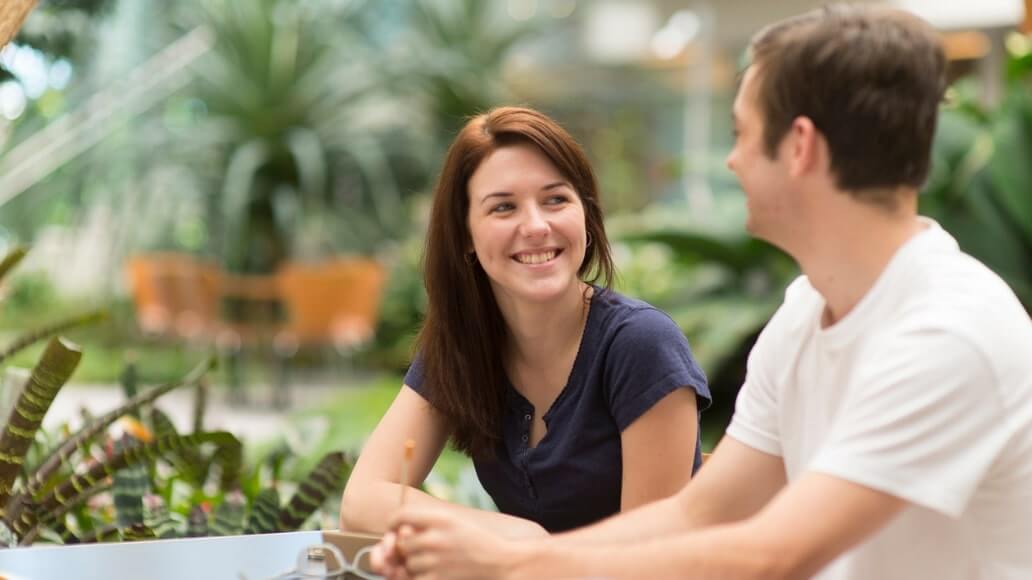 A woman and a man sit at a table outdoors, smiling and talking, surrounded by greenery.