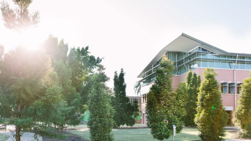 A modern building with large windows is surrounded by tall trees and greenery under a bright sky at UniSQ Ipswich campus.