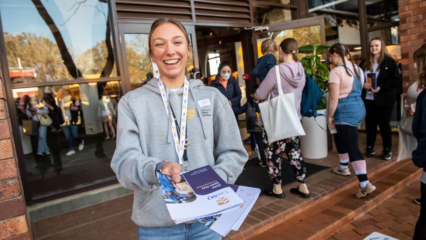 A smiling person hands out pamphlets to a group of people standing near the entrance of a building.