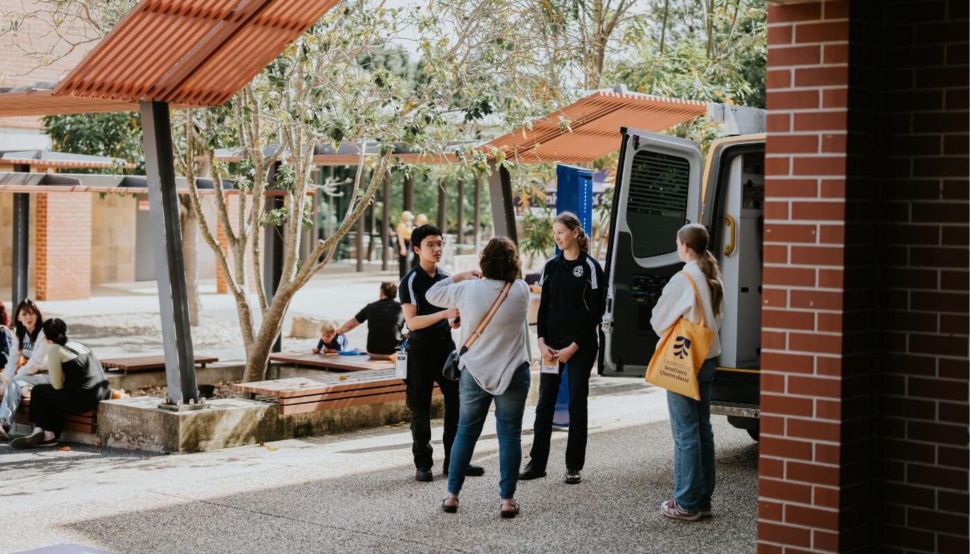 A group of people engages in conversation outside near a parked van, with several others sitting or walking nearby. Trees and brick structures can also be seen in the background.