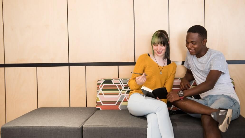 Two people sit on a sofa. One holds a notebook and a pencil, while the other points to the notebook, engaged in discussion.