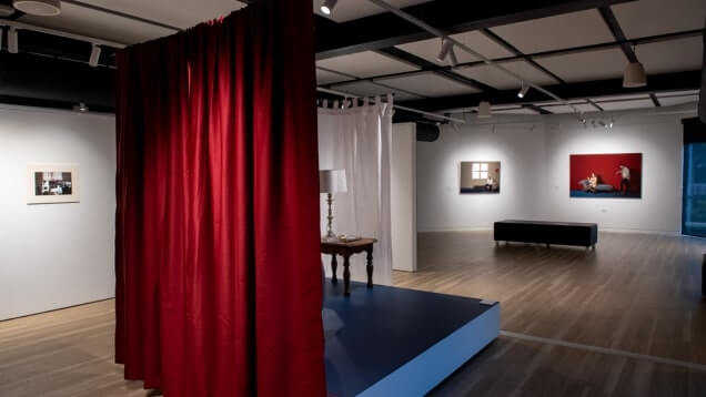 Art gallery interior with red curtains, framed artworks on white walls, wooden floor, and ambient ceiling lighting.