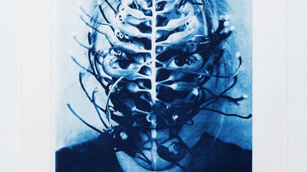 A surreal image of a person's face obscured by intricate, branching organic patterns, resembling a vine or root system. The artwork is in blue tones.