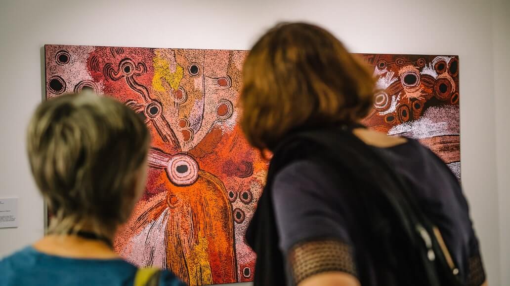 Two people observing an abstract painting with circular patterns and warm colors in an art gallery.