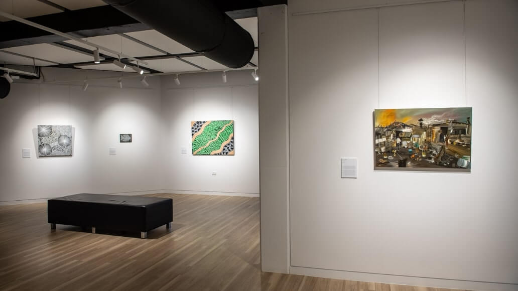 An art gallery room with paintings on white walls, wooden floor, and a black bench in the center.