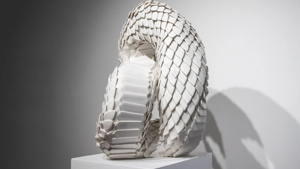 White geometric sculpture with interlocking, angular folds displayed on a pedestal in a gallery setting.