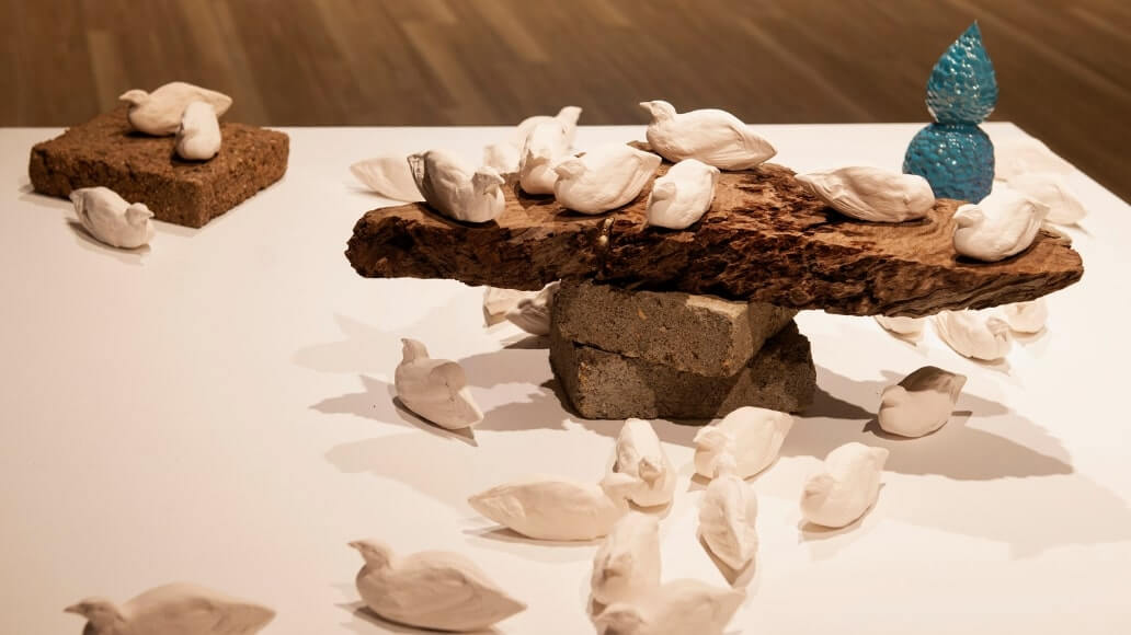 A group of white bird sculptures on and around a wooden slab supported by bricks. A blue textured sculpture is in the background.