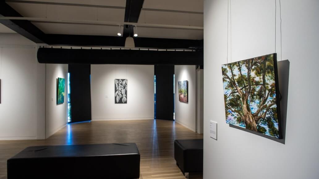 Art gallery interior with paintings on white walls, wooden floors, and black benches. A landscape painting with a tree is in the foreground.