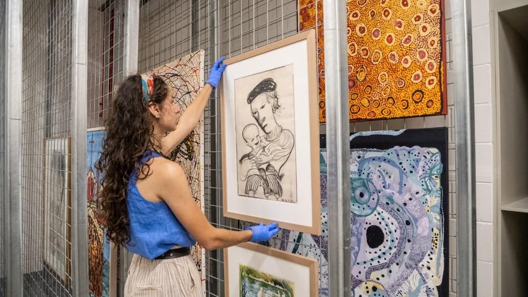 A person in a blue top and gloves hangs a framed drawing of a woman and child in an art storage area, surrounded by colorful abstract paintings.