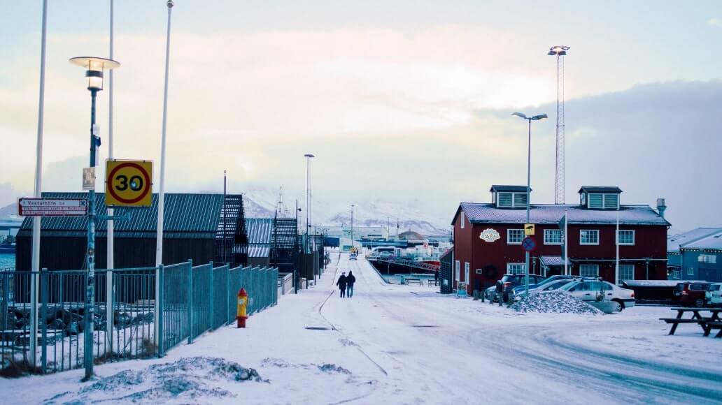 Snowy street scene with a 30 km/h sign, two people walking, industrial buildings on the left, and a red building on the right. Snow-covered mountains are visible in the background.