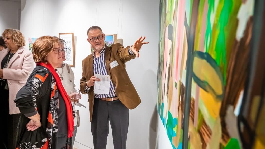 Two people discuss a large abstract painting at the UniSQ Art Gallery. Other patrons and artworks are visible in the background.